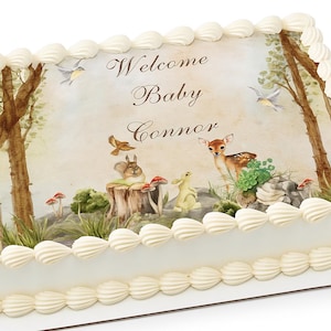 May include: A rectangular cake decorated with white frosting and a forest scene. The scene includes trees, a deer, a squirrel, a rabbit, and the text "Welcome Baby Connor". The cake is trimmed with white frosting.