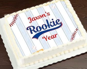 May include: A rectangular sheet cake with white frosting borders and a top surface featuring a light blue pinstripe pattern. The design includes red baseball stitching graphics, small baseballs, and the text Jaxon's Rookie Year in blue and red.