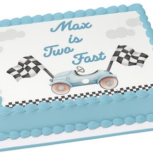 May include: A rectangular cake with light blue frosting and a white top. The top features a light blue race car, two checkered flags, and the text "Max is Two Fast". The cake is decorated with blue frosting around the edges.