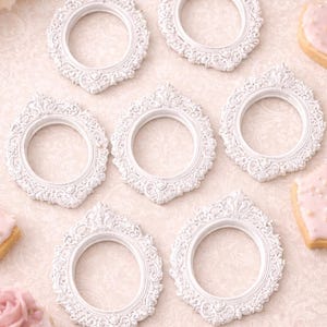 May include: Seven white, ornate, oval-shaped picture frames with detailed scrollwork are arranged on a patterned surface. The frames have a vintage, romantic aesthetic. Decorative cookies with pink frosting and flowers are also present.