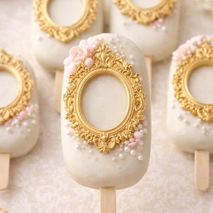 May include: White, oval-shaped cake pops on wooden sticks, decorated with gold ornate frames, pink flowers, and small white pearls. The cake pops are arranged on a white surface.