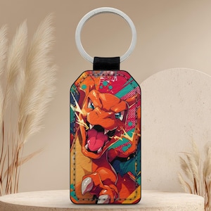 Könnte beinhalten: Ein rechteckiger Schlüsselanhänger mit einer Illustration von Charizard. Das Kunstwerk zeigt das Pokémon in Orange-, Rot- und Gelbtönen vor einem farbenfrohen Hintergrund. Der Schlüsselanhänger hat einen silbernen Ring und einen schwarzen Riemen. Maße in Zentimetern.