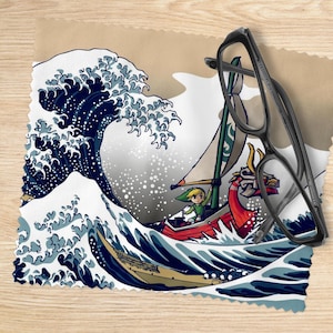 Pode incluir: Um pano de limpeza de microfibra com uma ilustração estilizada de um barco navegando por uma grande onda oceânica. O design inclui um personagem em um barco com uma cabeça de dragão vermelha. Um par de óculos pretos repousa sobre o pano.