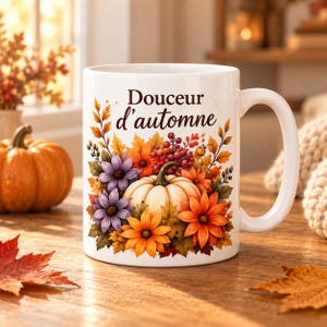 Peut inclure: Un mug en céramique blanche orné d'une illustration automnale colorée représentant une citrouille blanche centrale entourée de fleurs orange, jaunes et violettes, de baies rouges et de feuilles, avec le texte Douceur d'automne en brun.