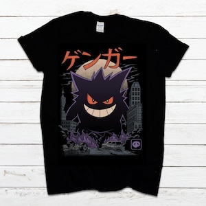 Peut inclure: T-shirt noir avec un motif de fantôme de dessin animé violet aux yeux rouges et un large sourire, sur fond de paysage urbain avec une pleine lune. Texte japonais au-dessus du fantôme. Le design utilise du violet et du gris.