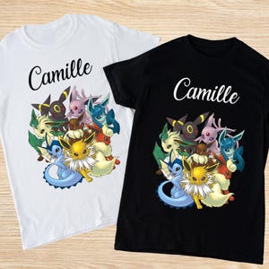 Puede incluir: Dos camisetas, una blanca y otra negra, con un gráfico de dibujos animados de varios personajes. El nombre "Camille" está impreso encima del gráfico en cursiva. El gráfico presenta personajes en amarillo, azul, rosa y verde.