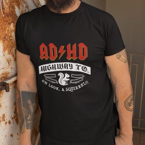 Può includere: T-shirt nera con la scritta "AD/HD Highway To... Oh Look, A Squirrel!!" in rosso e bianco. La maglietta presenta una grafica di scoiattolo ed è un'interpretazione umoristica del disturbo da deficit di attenzione e iperattività.