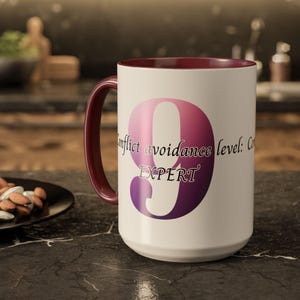 May include: A white ceramic mug with a burgundy interior and handle. The mug features a large, gradient pink and purple number 9 with the text "Conflict avoidance level: Expert". The mug is on a dark surface.