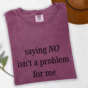 Könnte beinhalten: Ein pflaumenfarbenes Kurzarm-T-Shirt mit Rundhalsausschnitt. Das Shirt ist mit schwarzem Text bedruckt, der lautet: saying NO isn't a problem for me. Ein Etikett der Marke Comfort Colors ist an der Innenseite des Kragens sichtbar.