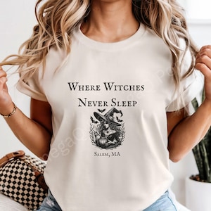 May include: A cream-coloured t-shirt featuring the text "Where Witches Never Sleep" above a black and white illustration of a witch with a hat and bats. The text "Salem, MA" is below the illustration.