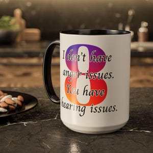 May include: White ceramic mug with a black handle and rim. The mug features the text "I don't have anger issues. You have hearing issues" in a gradient of purple, orange, and red. The mug is on a dark countertop.