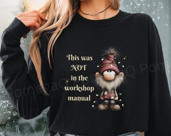 May include: Black long-sleeve sweatshirt with a cartoon gnome wearing a red hat, white beard, and fairy lights. The text "This was NOT in the workshop manual" is printed above the gnome. The sweatshirt is worn by a person.