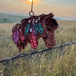May include: A brown horse-shaped ornament with colorful glitter accents and feather details. The ornament is hanging from a black string against a backdrop of a sunset over a grassy field. The horse has purple, teal, and red feathers.