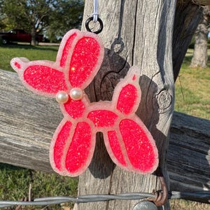 May include: A red glittery balloon dog-shaped air freshener with a light pink outline and two pearl beads. The air freshener hangs from a white string with a black ring. The background is a weathered wooden fence.
