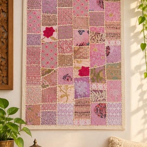 May include: A rectangular wall hanging featuring a patchwork design of various pink and purple fabric squares. The textile displays diverse floral patterns, intricate embroidery, and sequin embellishments, all joined together with visible white stitching.