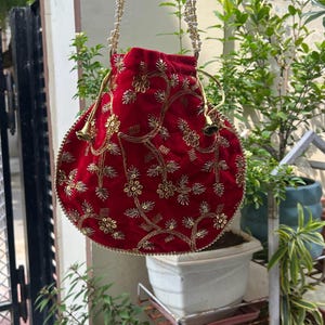 Puede incluir: Un bolso de mano de terciopelo rojo con bordados florales dorados y una correa de cuentas. El bolso redondo presenta un cierre de cordón y está adornado con ribetes dorados. Un accesorio decorativo.