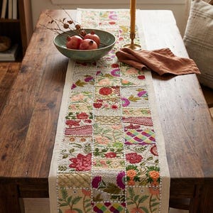 May include: A rectangular table runner featuring a patchwork design of embroidered floral and geometric patterns in vibrant colors including pink, green, orange, and red on a light beige fabric base with visible stitching and decorative beadwork.