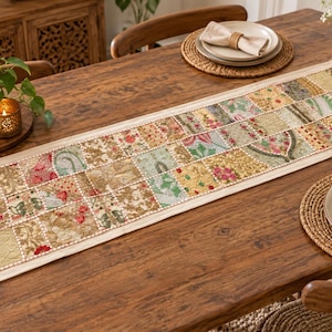 May include: A rectangular table runner featuring a patchwork design of various fabric squares. The squares display intricate floral and paisley patterns in shades of gold, green, red, and cream, bordered by a solid cream-colored fabric edge.