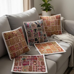 Puede incluir: Varios cojines decorativos con diseño de patchwork en tonos rojos, dorados y verdes, con bordes blancos. Están colocados sobre un sofá gris, creando un ambiente cálido y acogedor en un salón.