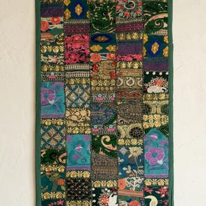 Puede incluir: Un tapiz rectangular que presenta un mosaico de varios cuadrados de tela en tonos verde, azul, morado y dorado. El tejido muestra bordados intrincados, motivos florales y detalles en hilo metálico, todo bordeado por un ribete de color verde.