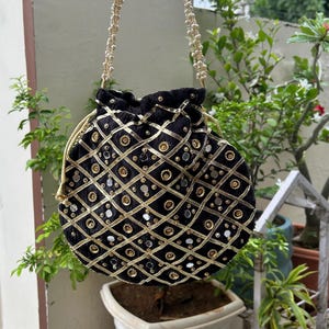 K&ouml;nnte beinhalten: Schwarzer Samtbeutel mit Kordelzug und goldenen und silbernen Verzierungen. Die Tasche hat ein Rautenmuster mit goldenen Verzierungen und kreisf&ouml;rmigen Akzenten. Der Riemen besteht aus kleinen, runden, goldenen Perlen.