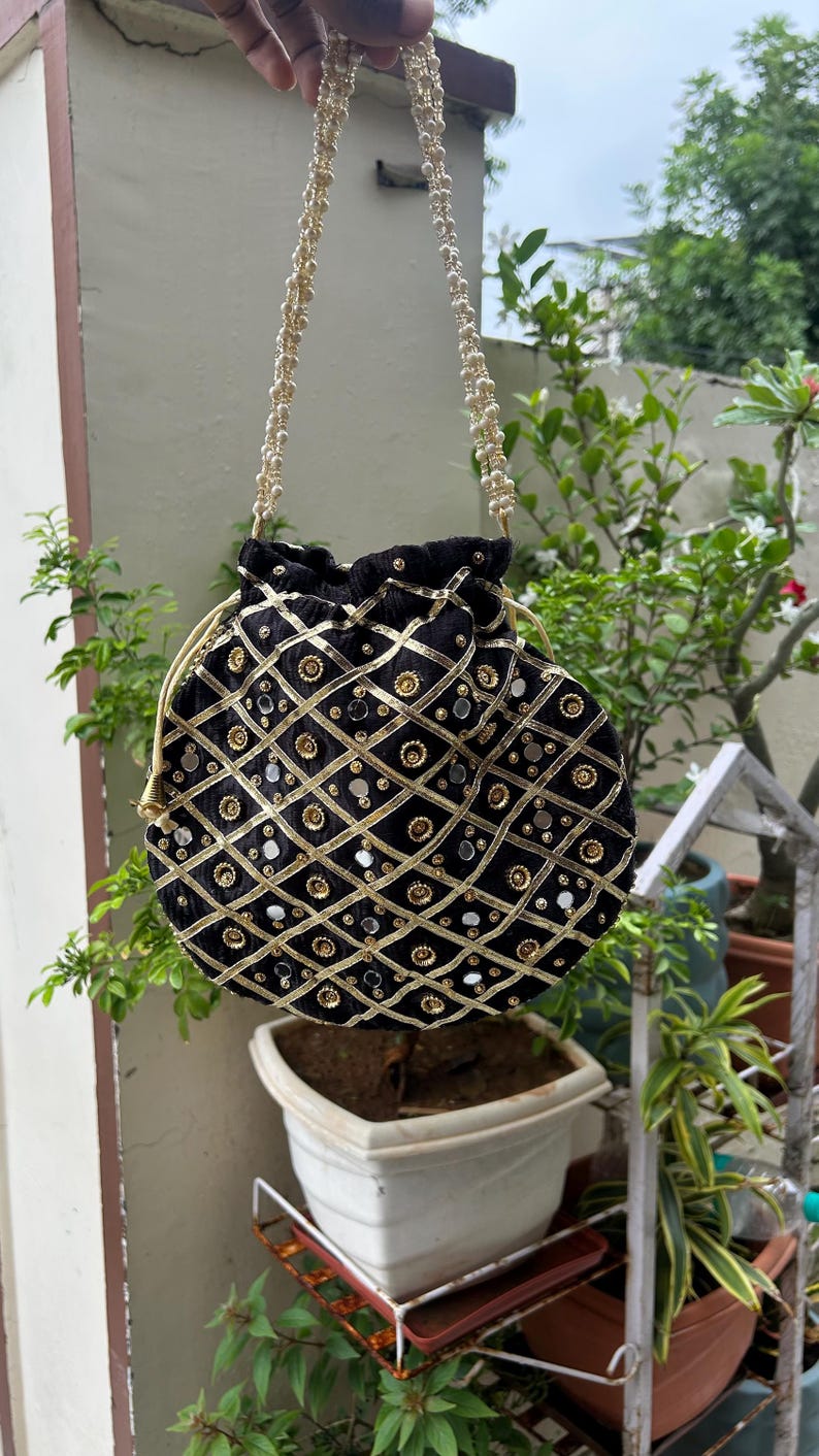K&ouml;nnte beinhalten: Schwarzer Samtbeutel mit Kordelzug und goldenen und silbernen Verzierungen. Die Tasche hat ein Rautenmuster mit goldenen Verzierungen und kreisf&ouml;rmigen Akzenten. Der Riemen besteht aus kleinen, runden, goldenen Perlen.