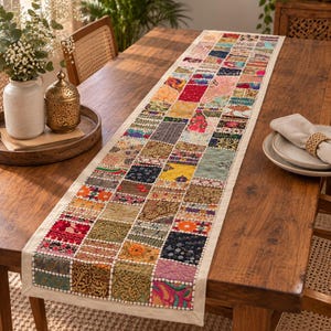 Può includere: Un lungo runner da tavola rettangolare caratterizzato da un design patchwork composto da vari quadrati di tessuto colorati. Ogni quadrato presenta motivi, ricami e perline distinti nei toni del rosso, blu, giallo, verde e oro, bordato da tessuto crema.