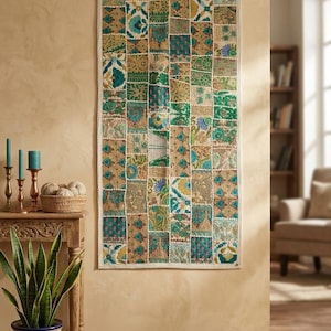 May include: A rectangular wall hanging featuring a patchwork design of various fabric squares in shades of teal, green, gold, and white. The textile displays intricate floral and geometric patterns with visible stitching and small white pom-pom accents.