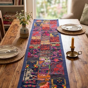 May include: A rectangular table runner featuring a patchwork design of dark blue fabric squares embroidered with colorful floral, leaf, and geometric patterns in shades of red, pink, orange, yellow, green, and white, finished with a solid blue border.