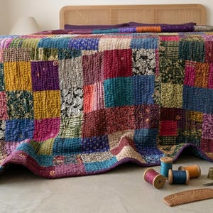 May include: A patchwork quilt in a variety of colors, including blue, yellow, pink, and purple, covers a bed. The quilt is made of squares of fabric sewn together. Sewing notions, including spools of thread, a thimble, and scissors, are on the floor.