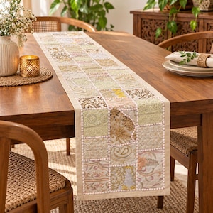 Puede incluir: Un camino de mesa rectangular con un diseño de patchwork de varios cuadrados de tela en tonos crema, beige y dorado suave. El camino incluye bordados intrincados, lentejuelas y cuentas, con un borde de color claro que enmarca el patrón.