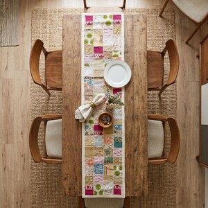 Peut inclure: Une table &agrave; manger rectangulaire en bois pr&eacute;sente un chemin de table en patchwork compos&eacute; de divers carr&eacute;s de tissu color&eacute;s. Une assiette blanche, une serviette pli&eacute;e avec un anneau dor&eacute;, un petit bol en bois et un vase sont pos&eacute;s dessus.