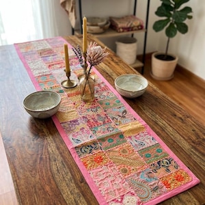 May include: A rectangular table runner featuring a patchwork design of various embroidered fabric squares in shades of pink, orange, teal, and gold. The runner has a solid pink border and is decorated with intricate floral and paisley patterns.