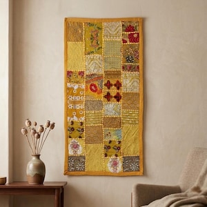 Könnte beinhalten: Ein rechteckiger Wandbehang mit einem Patchwork-Design aus verschiedenen gelben, goldenen und senffarbenen Stoffquadraten. Das Textil enthält aufwendige Stickereien, Pailletten und florale Muster, eingefasst von einem Rahmen aus einfarbigem gelbem Stoff.