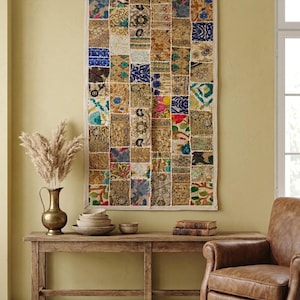 Könnte beinhalten: Ein rechteckiger Wandbehang mit einem Patchwork-Design aus verschiedenen Stoffquadraten in Gold-, Blau-, Grün- und Rosatönen. Das Textil zeigt komplizierte Muster, Stickereien und metallische Akzente, eingefasst von einem hellen Stoffrand.