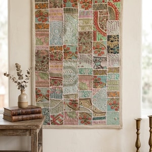 May include: A rectangular wall hanging featuring a patchwork design of various fabric squares in shades of green, pink, gold, and blue. The textile displays intricate floral embroidery, metallic thread accents, and white beaded trim details.