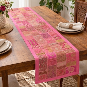 Peut inclure: Un chemin de table rectangulaire compos&eacute; d'un assemblage de carr&eacute;s de tissu rose. Chaque carr&eacute; pr&eacute;sente des broderies dor&eacute;es complexes, des accents de fils m&eacute;talliques et des motifs floraux ou cachemire, bord&eacute; d'un tissu rose uni.