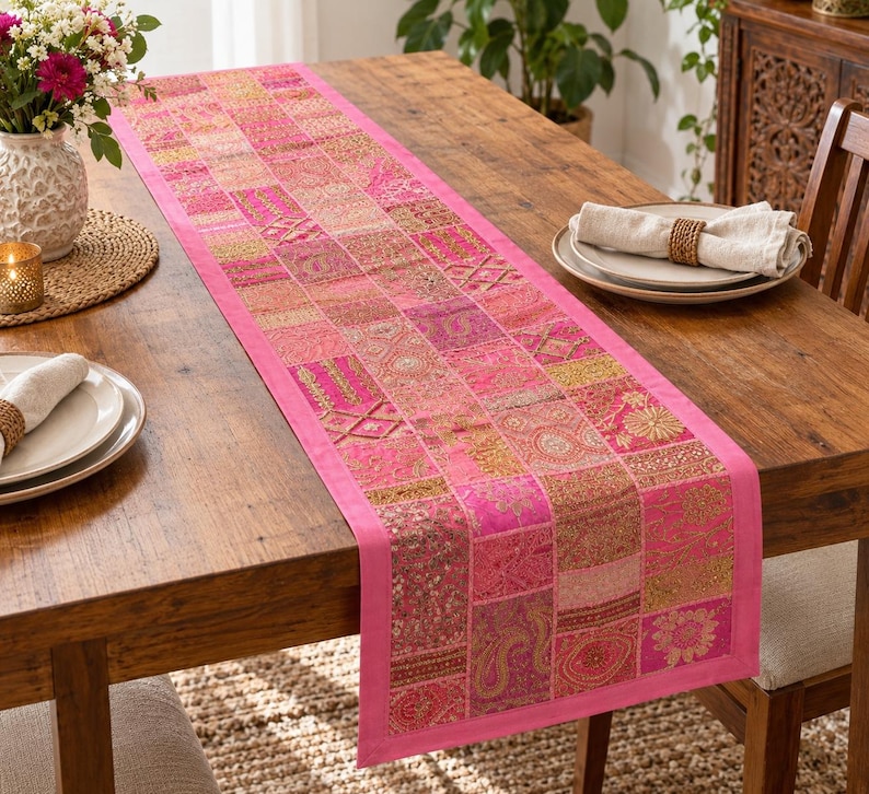 Peut inclure: Un chemin de table rectangulaire compos&eacute; d'un assemblage de carr&eacute;s de tissu rose. Chaque carr&eacute; pr&eacute;sente des broderies dor&eacute;es complexes, des accents de fils m&eacute;talliques et des motifs floraux ou cachemire, bord&eacute; d'un tissu rose uni.
