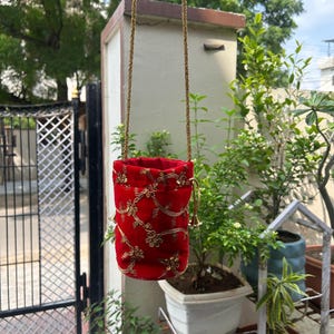 Może przedstawiać: Czerwona aksamitna torba ze sznurkiem, złotym haftem kwiatowym i złotym łańcuszkiem. Torba wisi przed ogrodem z zielonymi roślinami i metalową bramą.