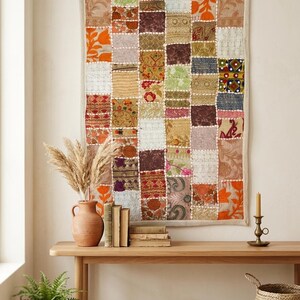 Puede incluir: Un tapiz rectangular con un diseño de patchwork compuesto por varios cuadrados de tela en tonos naranja, marrón, verde y crema. El tejido muestra diversos patrones florales y geométricos, con costuras blancas visibles uniendo cada cuadrado.