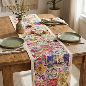 May include: A rectangular table runner featuring a patchwork design of embroidered floral and geometric patterns in vibrant shades of pink, yellow, purple, and gold on a cream-colored fabric base, laid flat on a wooden dining table with place settings.