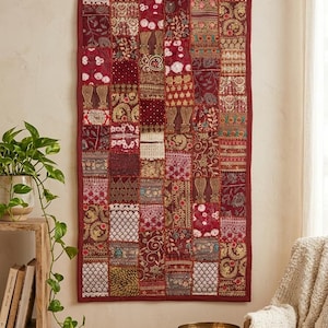 Op de afbeelding: Een rechthoekig wandkleed met een patchworkontwerp van diverse stoffen vierkanten in tinten dieprood, bordeaux en goud. Het textiel toont ingewikkeld borduurwerk, accenten van metaaldraad en diverse bloemrijke en geometrische patronen.