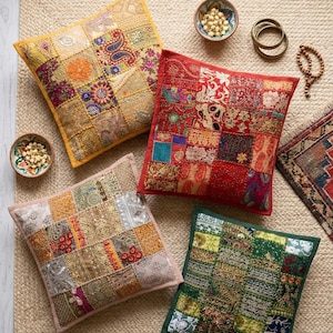 Puede incluir: Cuatro cojines cuadrados decorativos con diseños de patchwork en tonos dorados, rojos, rosas y verdes. Los cojines están colocados sobre una alfombra tejida, con pequeños cuencos de cuentas y pulseras cerca. Estilo bohemio.