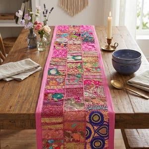 Peut inclure: Un chemin de table rectangulaire composé d'un assemblage de carrés de tissu brodés dans des tons de rose, magenta, or et bleu. Le chemin de table présente une bordure rose unie et des détails complexes en fil métallique sur les motifs.