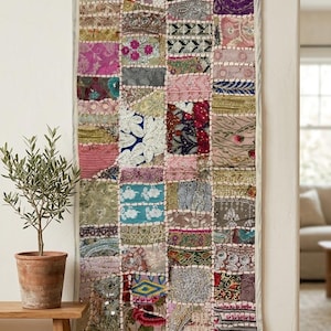 May include: A patchwork wall hanging with a multitude of fabric squares in various colours and patterns. The squares include floral and geometric designs in pink, blue, green, and purple. The edges are framed with a light-coloured border.