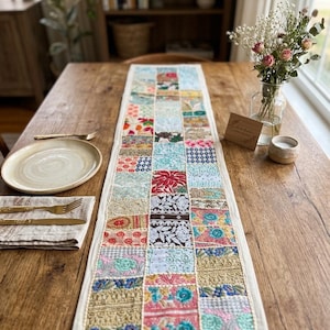 May include: A rectangular table runner featuring a patchwork design of various embroidered fabric squares in vibrant colors including red, blue, gold, and white. The runner has a cream-colored border and is centered on a wooden dining table.