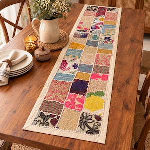 May include: A rectangular table runner featuring a patchwork design of various fabric squares in colors including pink, yellow, blue, green, and beige. The runner has a solid cream-colored border and visible white stitching between the fabric patches.