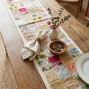 Peut inclure: Un long chemin de table rectangulaire compos&eacute; d'un patchwork de carr&eacute;s de tissu brod&eacute;s dans des tons de rose, vert, dor&eacute; et bleu. Le chemin de table est agr&eacute;ment&eacute; d'une serviette cr&egrave;me dans un rond de serviette dor&eacute;, d'un vase et d'un bol en bois.