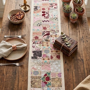 May include: A patchwork table runner with a cream border, featuring squares of patterned fabrics in pink, red, and blue. The runner is on a rustic wooden table, with a bowl, plates, and small potted plants. The table runner is approximately 150 cm long.
