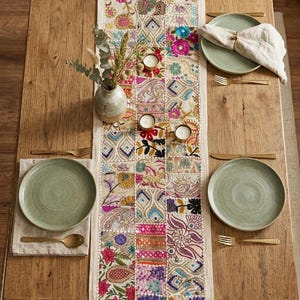 May include: A rectangular table runner featuring a patchwork design of embroidered fabric squares in vibrant colors including pink, blue, yellow, and green. The runner is centered on a wooden table with four green plates, gold cutlery, and candles.